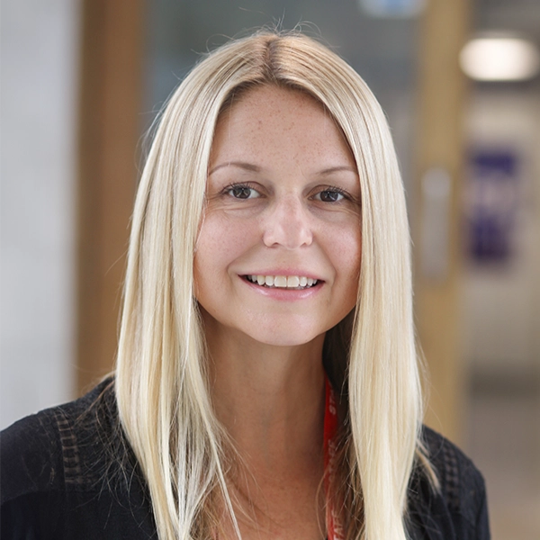 A person with long, straight light‑coloured hair is shown from the shoulders up in an indoor setting with soft, neutral lighting and blurred background features.