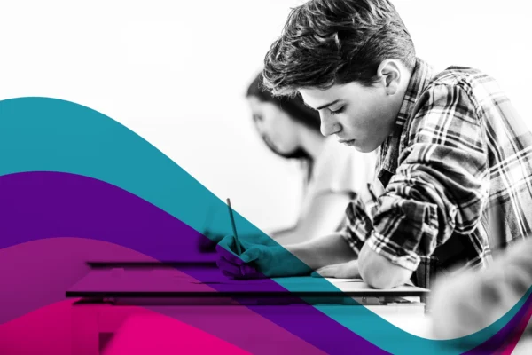 A person seated at a desk writing on paper in a classroom environment. A flowing graphic in teal, purple, and pink curves across the lower part of the image.