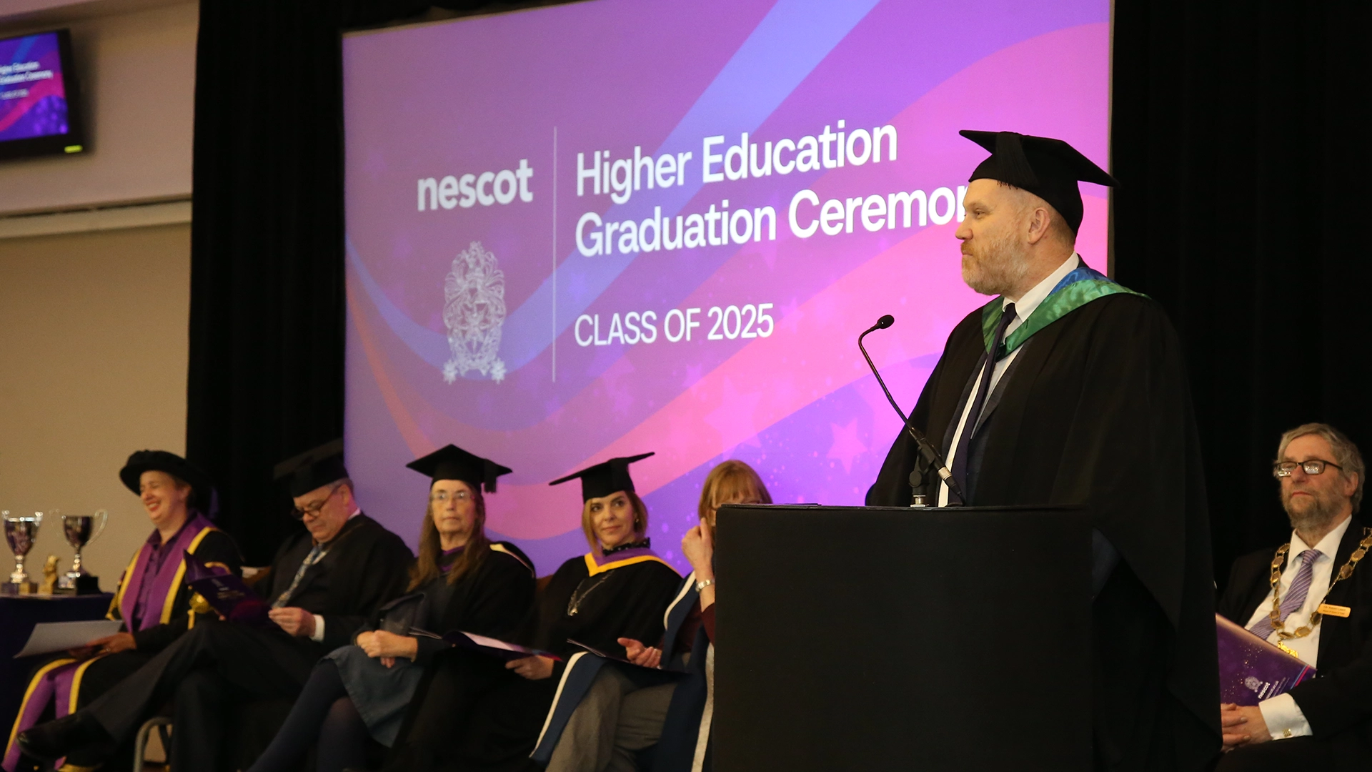 A graduation ceremony stage with graduates seated in academic gowns while a speaker in a gown and cap stands at a lectern. A large screen behind them displays the text “nescot Higher Education Graduation Ceremony, Class of 2025.”