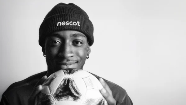 A person wearing a knitted hat with the word “nescot” on the front is holding a football close to their chest against a plain background.