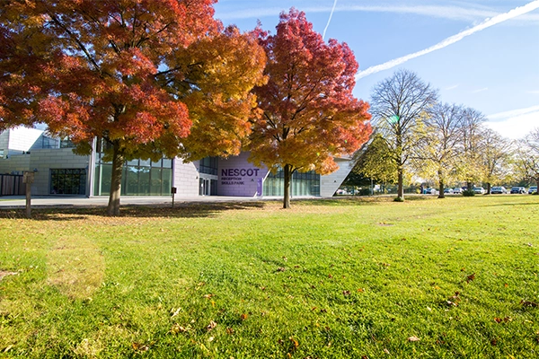 A grassy lawn in front of the Nescot campus with large trees displaying bright orange and red autumn foliage and a purple Nescot banner visible on the building.