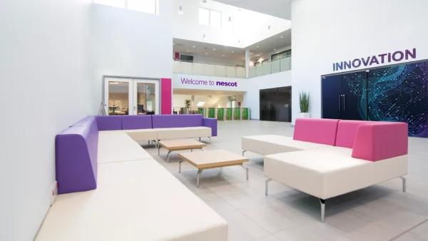 A spacious lobby area with purple, white, and pink modular seating arranged around low wooden tables. Visible signage includes “Welcome to nescot” and “INNOVATION” on the surrounding walls.