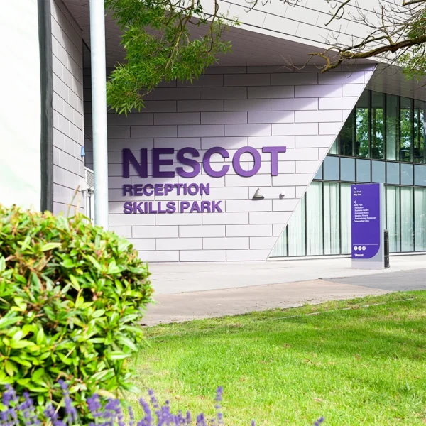 The exterior of the Nescot Skills Park building with large purple signage reading “NESCOT Reception Skills Park.” The scene includes landscaped greenery and modern angular architecture.