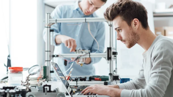Two people working in a lab; one person uses a laptop while another adjusts equipment on a 3D printer.