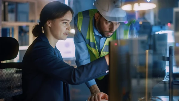 Two people in an industrial workspace reviewing information on a computer, with one wearing a safety vest and hard hat.