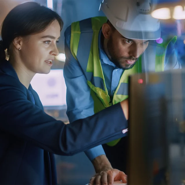 Two people in an industrial workspace reviewing information on a computer, with one wearing a safety vest and hard hat.