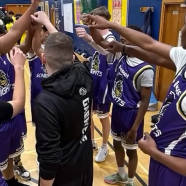 Group of basketball players in matching Nescot Knights jerseys standing in a circle on an indoor court with arms raised in a team huddle.