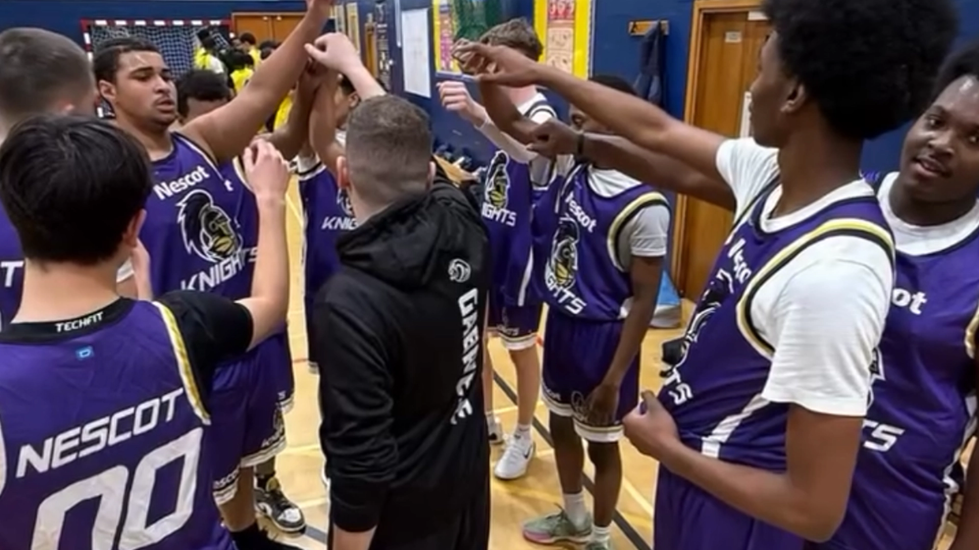 Group of basketball players in matching Nescot Knights jerseys standing in a circle on an indoor court with arms raised in a team huddle.