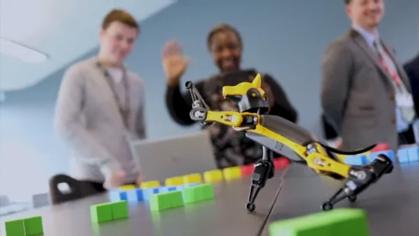 A small yellow robotic dog positioned on a table among colourful square blocks, with three people standing in the background observing the demonstration.