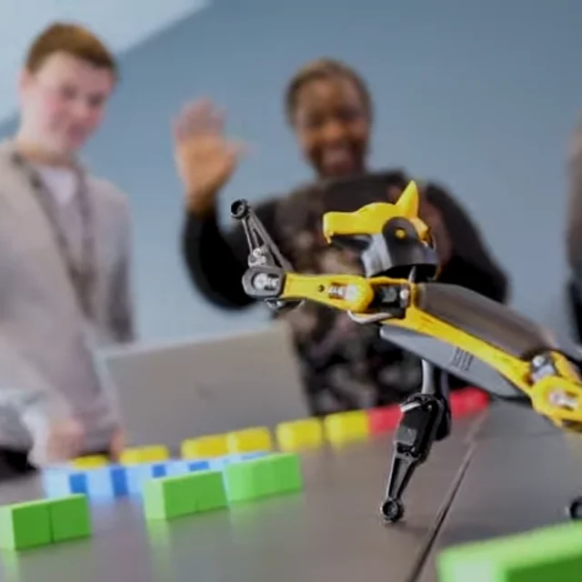 A small yellow robotic dog positioned on a table among colourful square blocks, with three people standing in the background observing the demonstration.