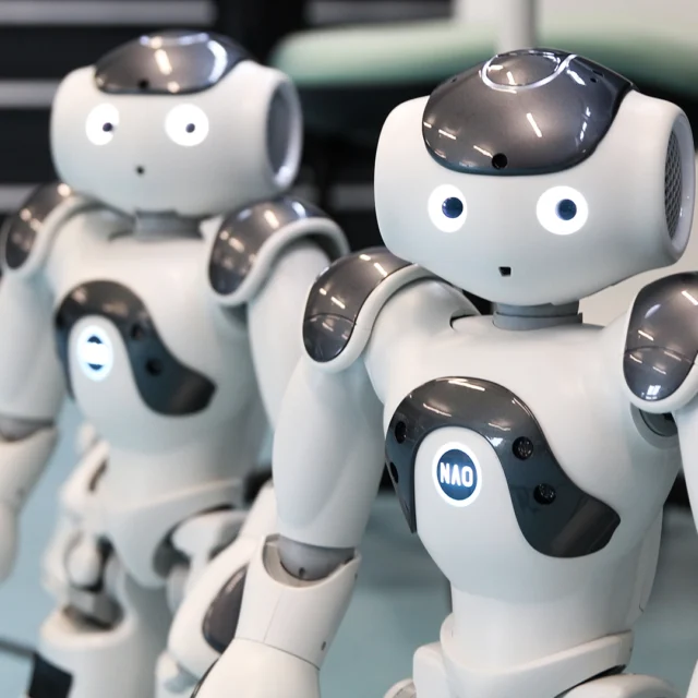 Two small humanoid robots with white and grey casing standing on a light blue floor in a technology lab, each with illuminated circular eyes and chest panels labelled “NAO.”