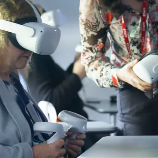 A person seated in a classroom wearing a virtual reality headset and holding two VR controllers while another person stands nearby holding an additional headset.