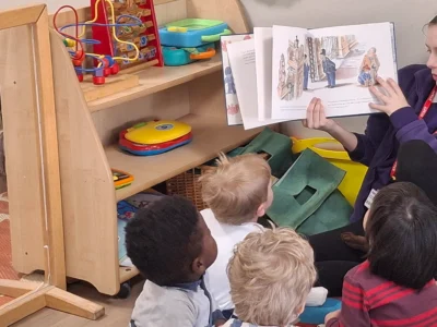 A young adult reads a picture book to a small group of children seated on the floor in a classroom with toys and shelves in the background.