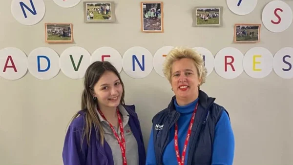 Two adults wearing lanyards stand indoors in front of a wall display with circular letters and small photos arranged in a row.