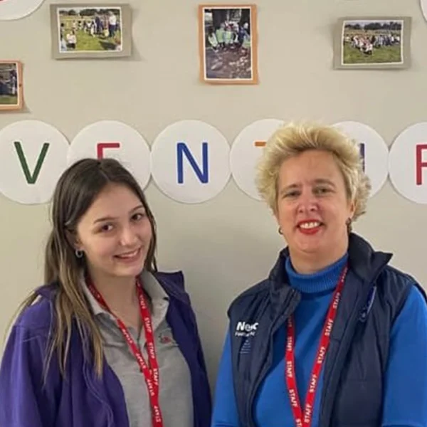 Two adults wearing lanyards stand indoors in front of a wall display with circular letters and small photos arranged in a row.