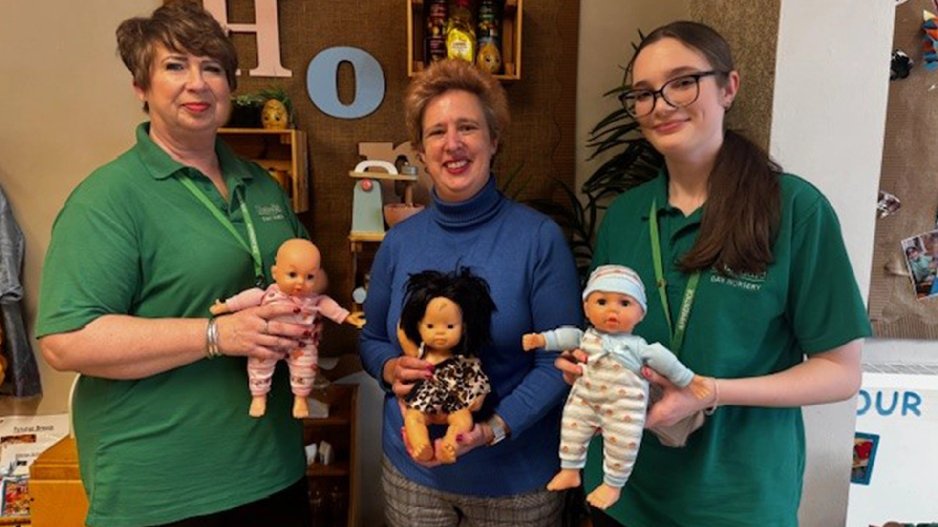 Three adults stand indoors holding toy dolls, with a decorated wall and shelves of children’s items in the background.