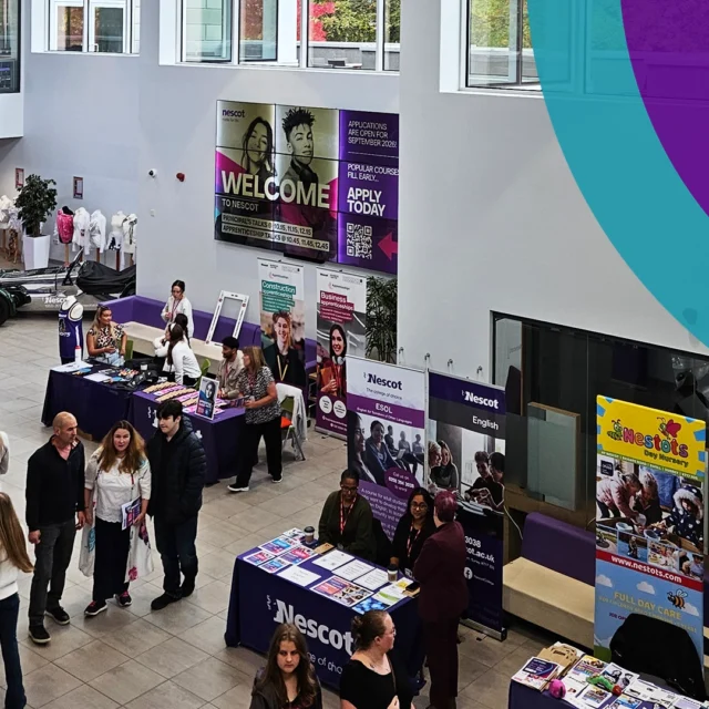 An overhead view of a college open event in a spacious atrium, showing several tables with course information, each covered with purple cloths and branded displays. Many visitors walk around the area, stopping to speak with staff at different stands. Large welcome posters and course advertisements are visible on the walls, with natural light entering through upper‑level windows.