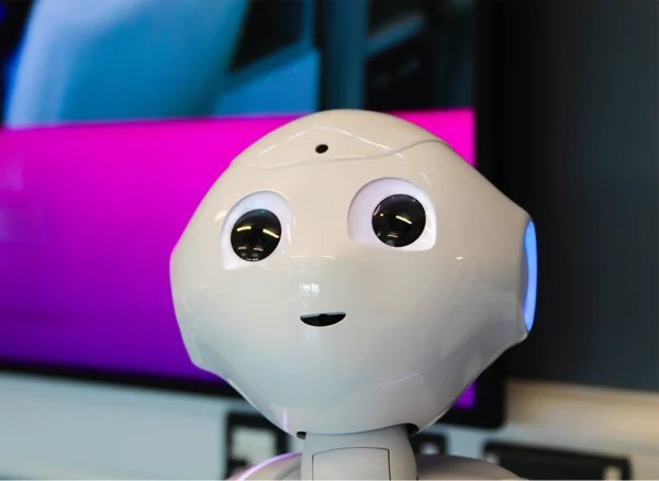 A close‑up view of a white humanoid robot with rounded features and large illuminated eyes, positioned in front of a screen displaying bright colours.