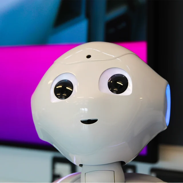 A close‑up view of a white humanoid robot with rounded features and large illuminated eyes, positioned in front of a screen displaying bright colours.