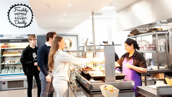 Several people stand in the refectory queue while a catering staff member serves hot food from behind the counter. Refrigerated grab‑and‑go items are visible in the background, along with a sign that reads “Freshly prepared in our kitchen daily.”