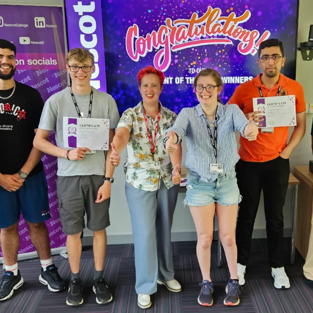 Group of eight people standing indoors in front of a screen that reads ‘Congratulations!’ Several individuals hold certificates, and promotional banners are visible behind them
