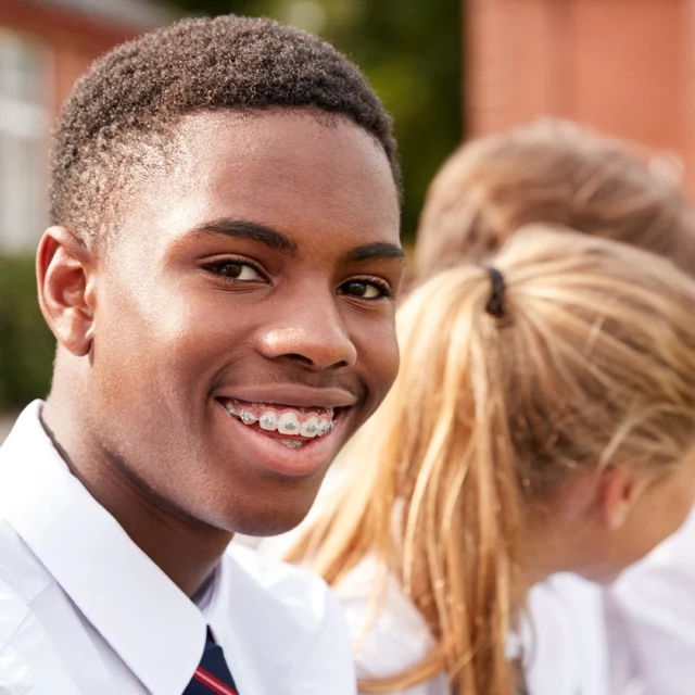 A row of students in school uniforms sitting outdoors near a brick building, viewed from the side, with one student in the foreground and others seated beside them.