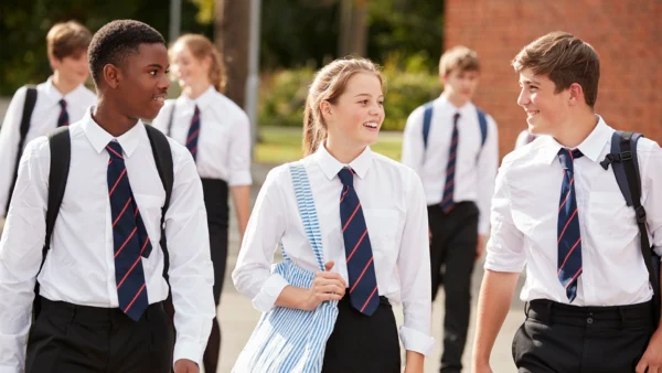 A group of students in school uniforms walking together outside on a pathway, carrying backpacks with a brick building and greenery in the background.