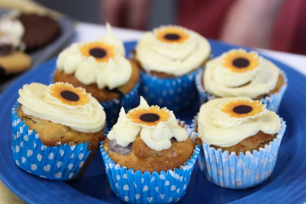 A plate of cupcakes in blue patterned wrappers, each topped with cream frosting and a sunflower‑style edible decoration.
