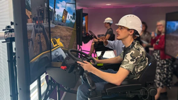An indoor training setup where a person wearing a safety helmet operates a construction vehicle simulator, using a seated console with steering wheel and multiple screens displaying the virtual environment.