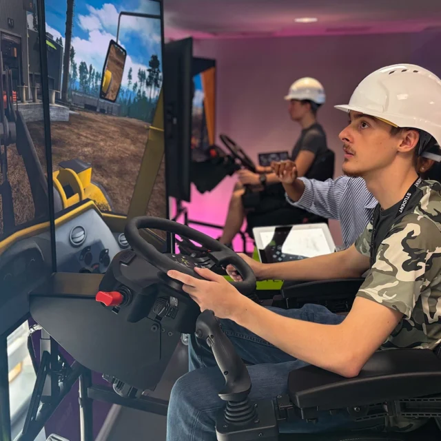 An indoor training setup where a person wearing a safety helmet operates a construction vehicle simulator, using a seated console with steering wheel and multiple screens displaying the virtual environment.