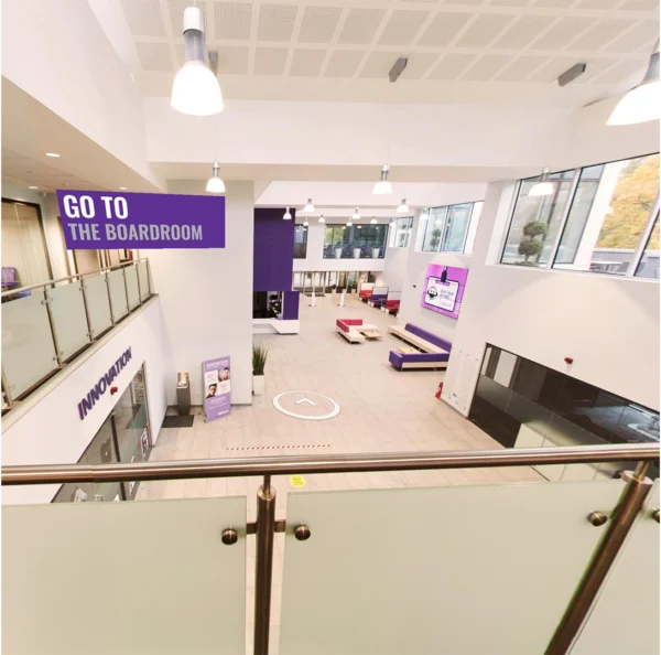 A wide view of a bright, modern atrium with white walls, large windows, and hanging ceiling lights. The space includes seating areas in purple and white, display screens, and signage pointing toward the boardroom. A glass balcony overlooks the ground floor.