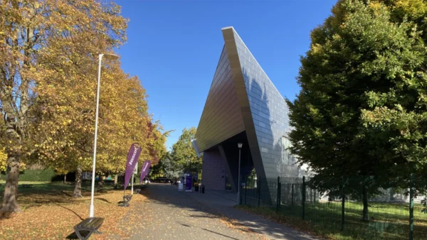 A modern angular building with a metallic exterior situated beside a tree‑lined walkway. Autumn leaves cover parts of the ground, and purple campus flags line the path.