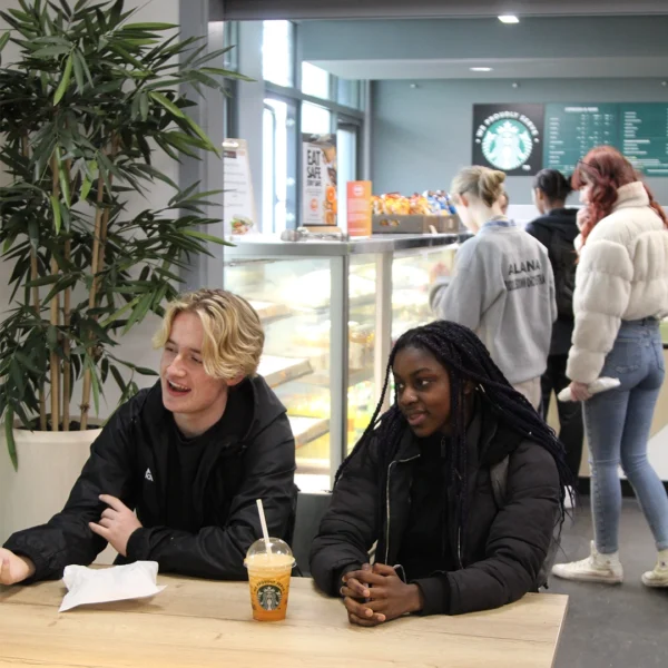 Two students seated at a table in a campus café, with a display counter of food and drinks behind them and other customers queuing near the counter.