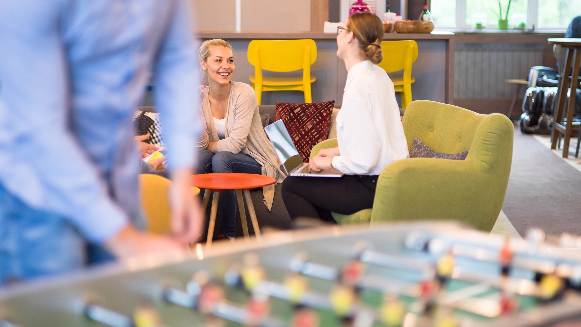 People sitting and talking in a brightly furnished common room, with a foosball table blurred in the foreground.