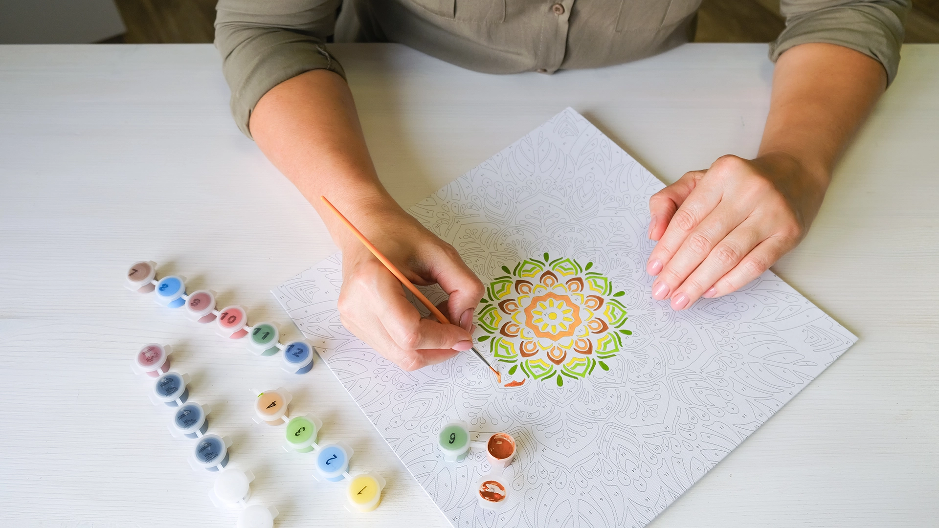 Person seated at a table using a paintbrush to fill in a detailed mandala-style colouring sheet, with small paint pots arranged beside the artwork.