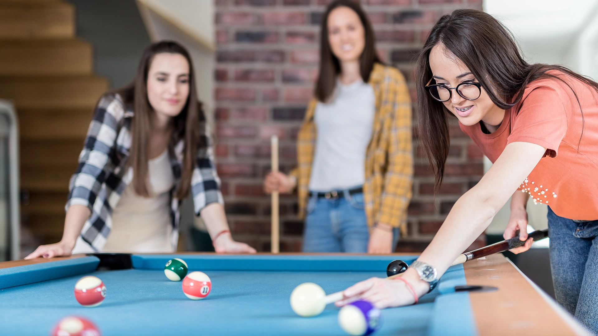 Three people standing around a pool table, with one person taking a shot while the others watch.