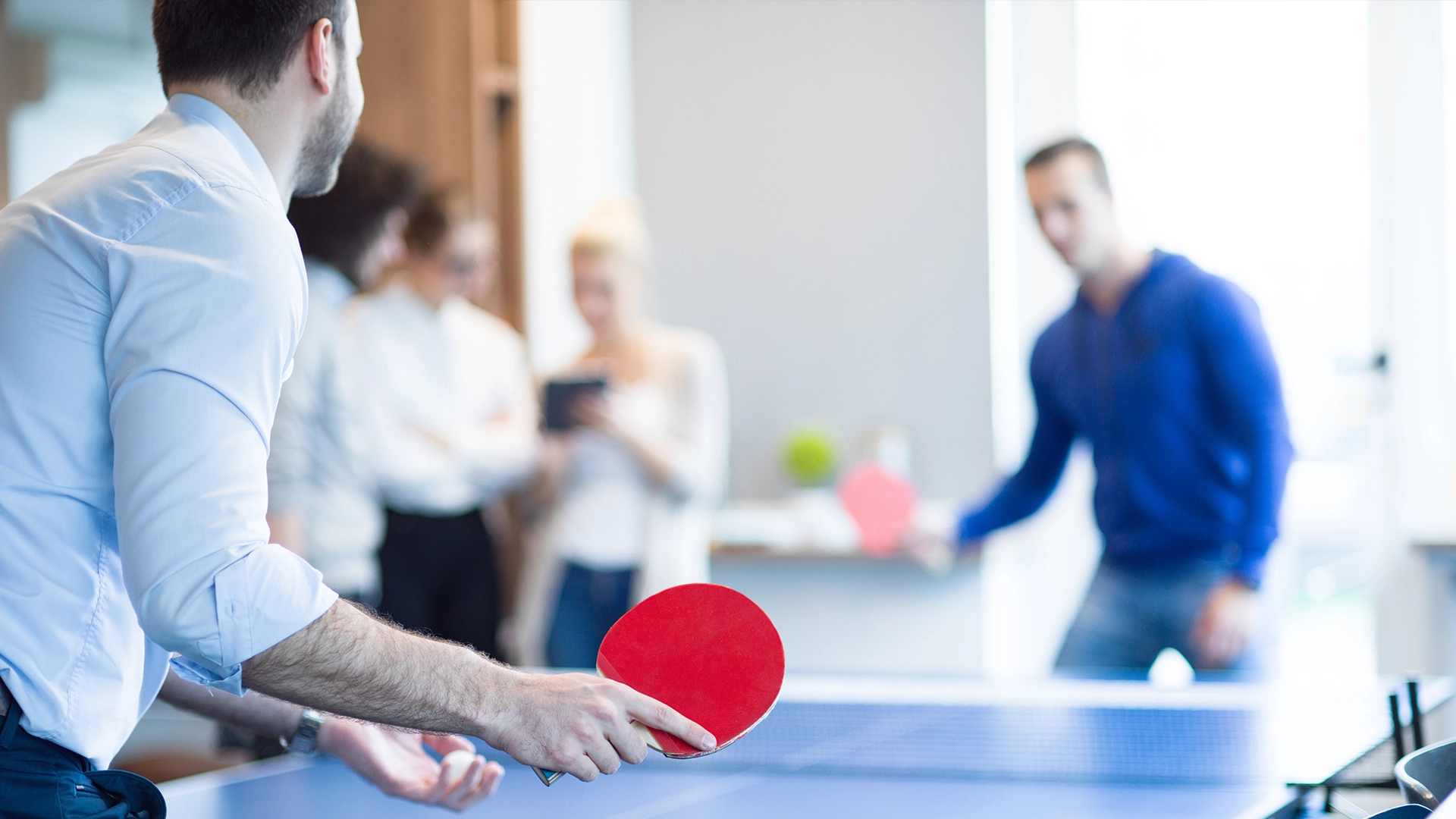 Two people playing table tennis indoors, with others standing and talking in the background.