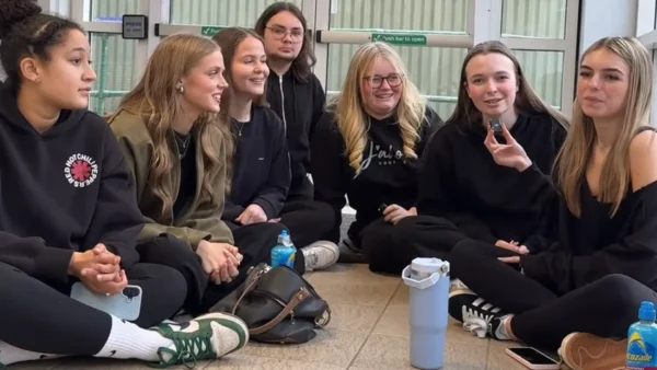 A group of seven students sit in a circle on the floor inside a building, talking together. Bags and water bottles are on the floor around them, with glass doors in the background.