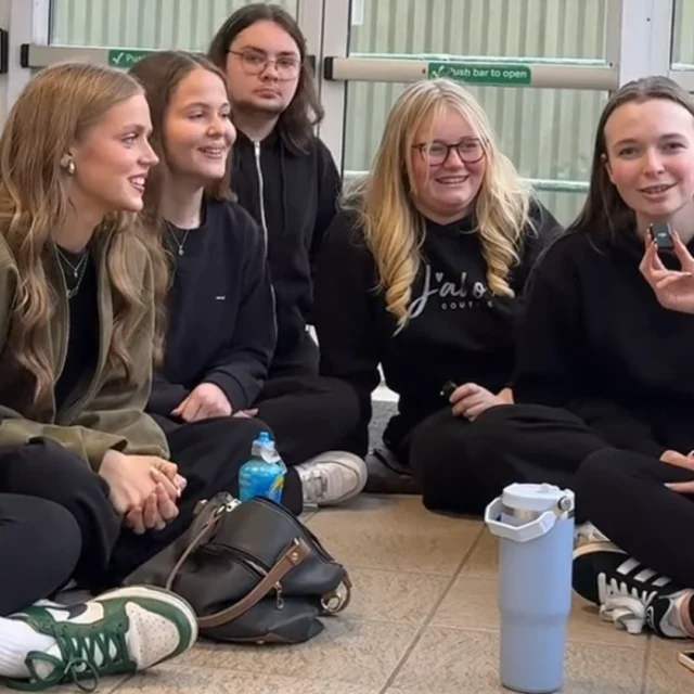 A group of seven students sit in a circle on the floor inside a building, talking together. Bags and water bottles are on the floor around them, with glass doors in the background.
