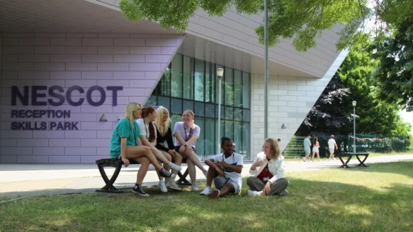 A group of students sitting on the grass outside the Nescot Skills Park building, which features large glass windows and purple signage. Trees and a walkway with people passing by are visible in the background.