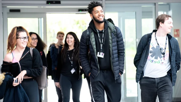 A group of students wearing lanyards walk through a set of glass entrance doors inside a campus building. Some students carry bags or jackets, and the background shows the bright outdoor light coming through the doorway.