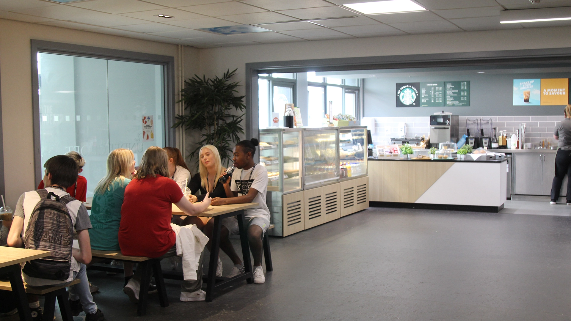 A group of students seated around tables inside a campus café area, with refrigerated food displays and a counter visible in the background.
