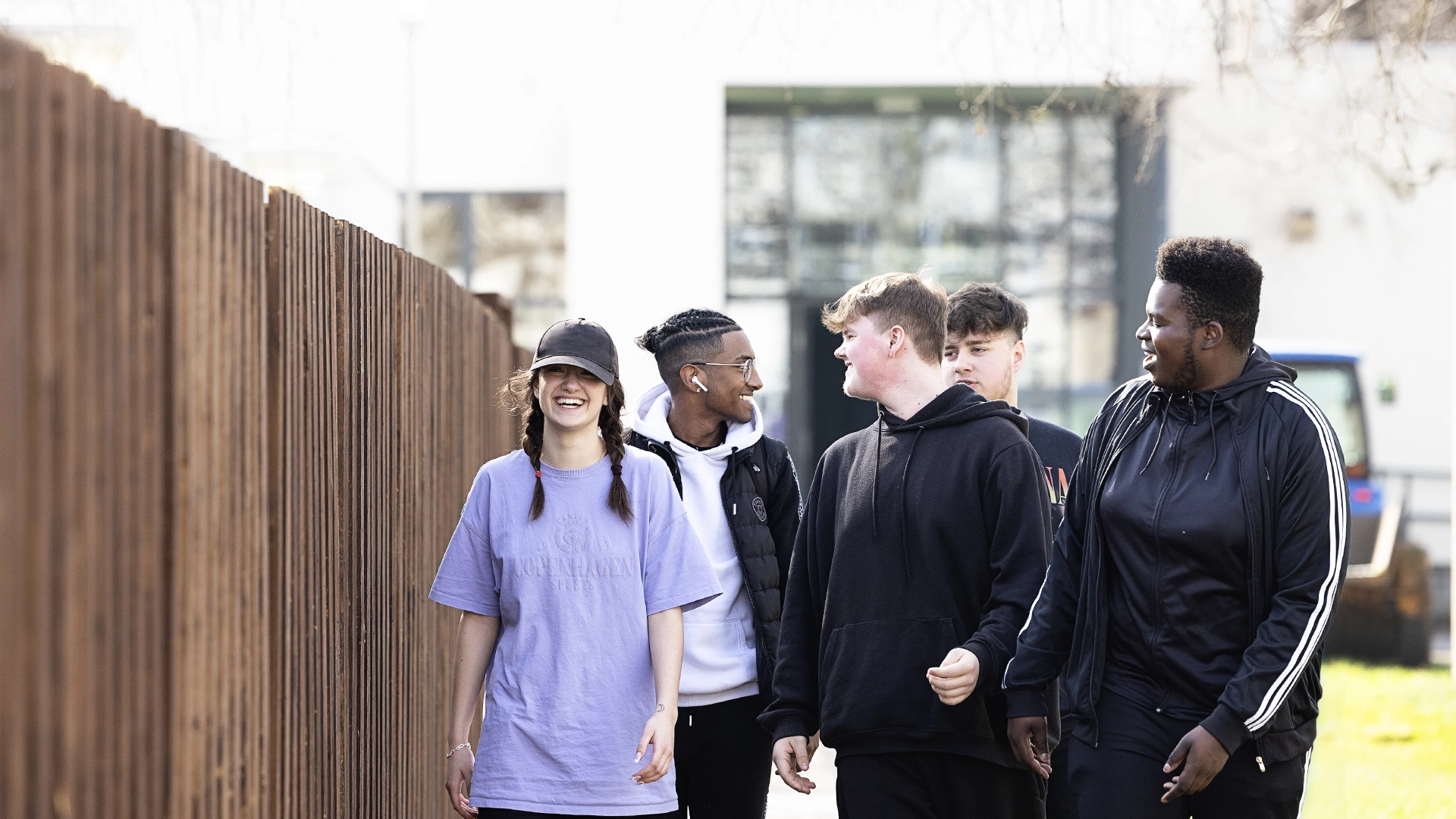 A group of students walk together along an outdoor pathway beside a tall wooden fence, with campus buildings and trees visible in the background.