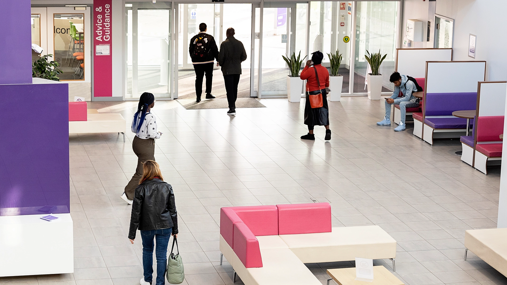 Several students walk through a spacious campus atrium with tiled flooring, bright lighting, and pink‑and‑purple seating areas. A sign for “Advice & Guidance” is visible near the entrance, and some students are seated along the side benches.