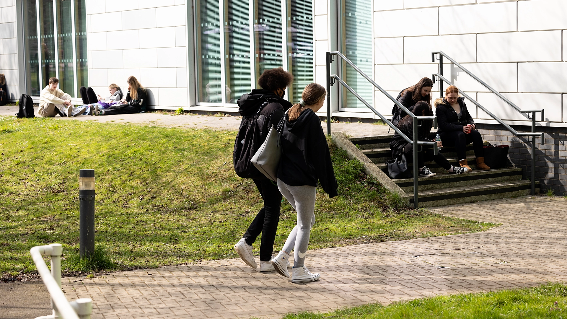 Students walk along a paved pathway beside a modern campus building with large windows, while others sit on the nearby steps and grassy bank. Trees and landscaped areas surround the open space.
