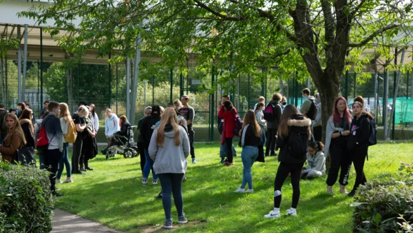Students gather in a grassy outdoor space with trees and fencing, standing and sitting in small groups across the lawn.
