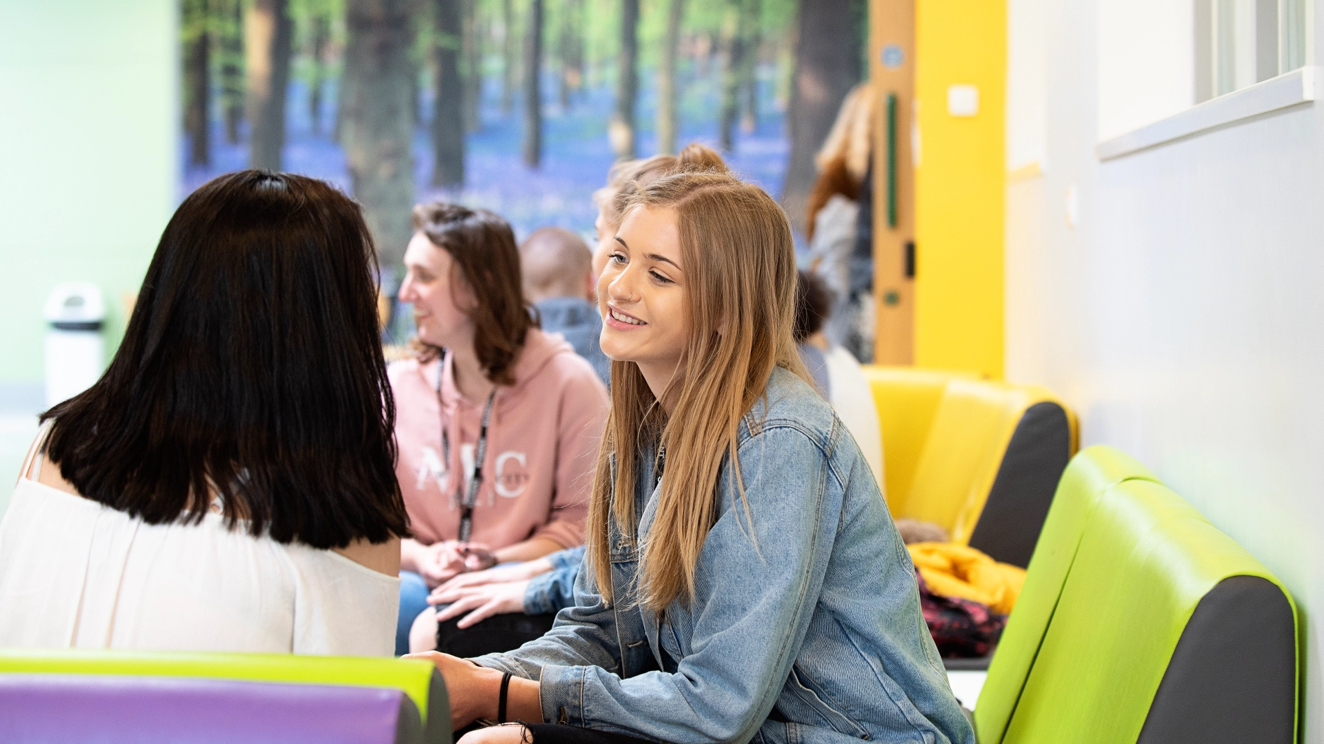 Several students sit on brightly coloured seating in an indoor common area, with green and purple cushioned benches and a wall mural of a forest scene in the background.