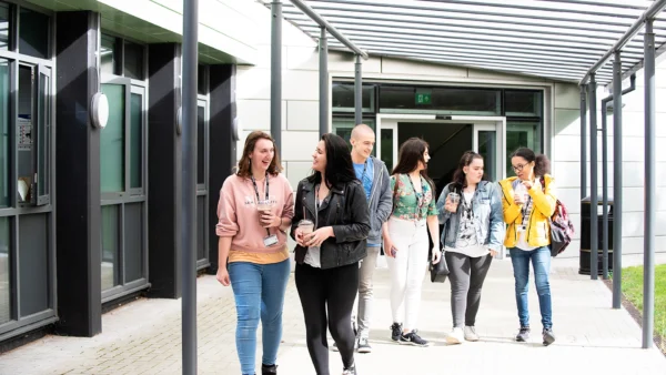 A group of students walk together along a covered walkway outside a campus building, holding drinks and bags as they move away from the entrance.