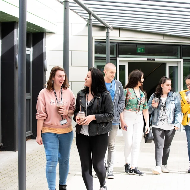 A group of students walk together along a covered walkway outside a campus building, holding drinks and bags as they move away from the entrance.