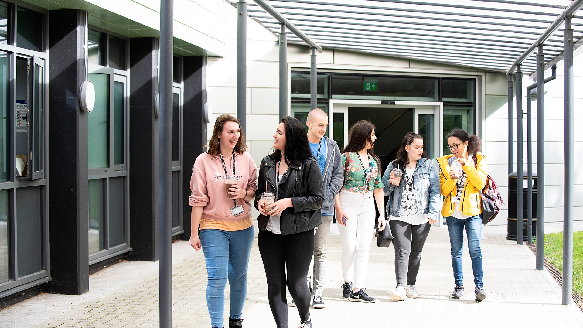 A group of students walk together along a covered walkway outside a campus building, holding drinks and bags as they move away from the entrance.
